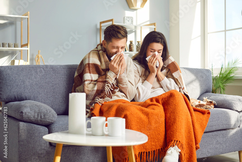 Young couple blowing their nose in tissue at home. Freezing man and woman covered with plaids sitting on sofa in living room having runny nose. Sick couple got flu, catch cold