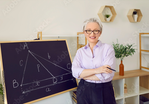 Maths teacher in front of blackboard in school classroom. Happy beautiful elegant middle aged woman in shirt and glasses standing by chalk board with geometry theorem, looking at camera and smiling