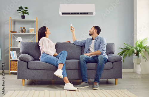 Young happy man and woman turning on air conditioner sitting on sofa at home. Smiling couple of homeowners enjoying cool conditioned air using remote resting on couch together in living room.