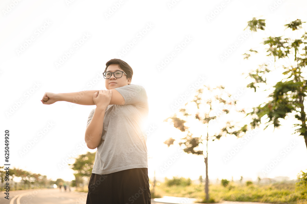 Young Asian man stretching body warm up before workout at outdoor with trees and light sunset background. Man exercises outside for health and wellbeing. Workout Exercising concept.