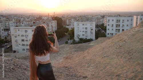 Sexy girl taking photos and videos of an African city with her phone in the sunset from a hill