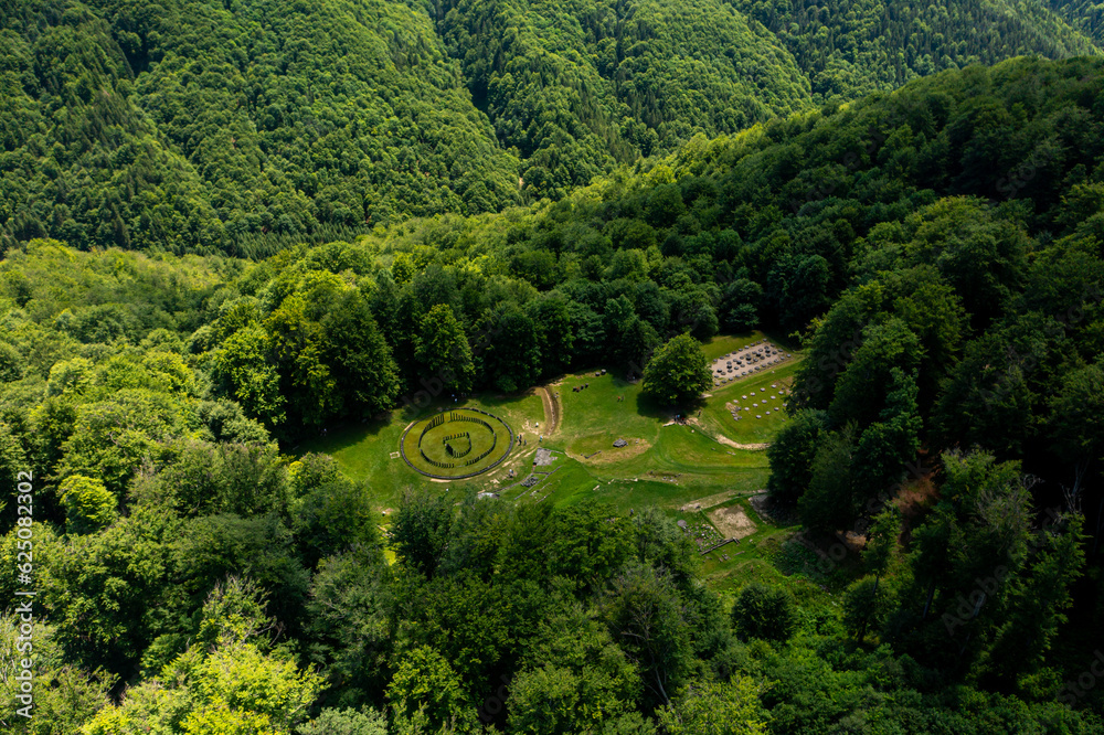 Aerial view over Sarmizegetusa Fortress in Romania. Photo from above during a beautiful sunny summer day. This historical fortress is surrounded by forests in Orastiei Mountains. Romania landmark. Aerial view over Sarmizegetusa Fortress in Romania. Photo from above during a beautiful sunny summer day. This historical fortress is surrounded by forests in Orastiei Mountains. Romania landmark.