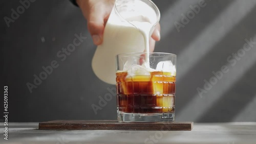 Male hand pouring whipped milk in to a glass with espresso coffee and ice cubes, process of making cold iced latte coffee