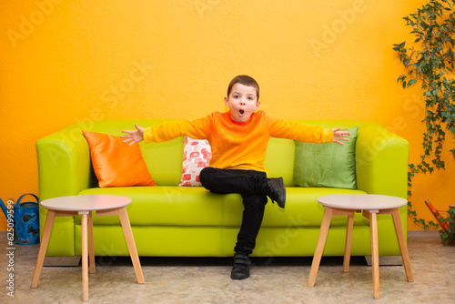 Surprised six-year-old schoolboy child in a bright orange sweater sits on a green sofa with his arms outstretched to the sides