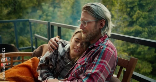 A girl hugs a blond guy with glasses. A couple is resting on a devan wrapped in a blanket on the balcony of a country house overlooking the mountains and forest photo