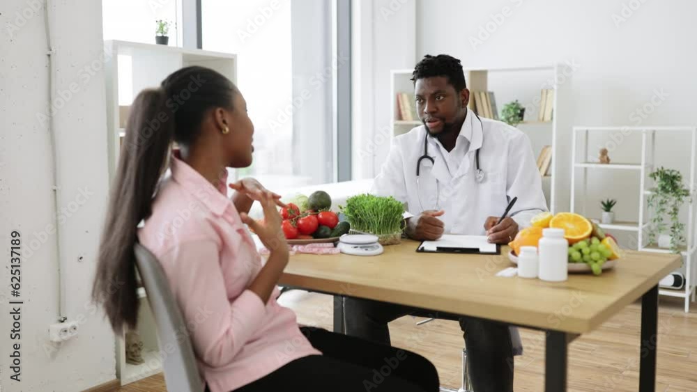 Selective focus of multiethnic man in lab coat sitting at desk with woman in consulting room of hospital. Experienced specialist in nutrition giving recommendations about healthy food choices. Selective focus of multiethnic man in lab coat sitting at desk with woman in consulting room of hospital. Experienced specialist in nutrition giving recommendations about healthy food choices.