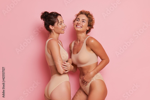 Two female models dressed in beige underwear posing on pink background, talking and laughing, happy friends concept, copy space