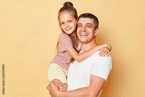 Positive optimistic father and daughter wearing casual t-shirts standing isolated over beige background dad holding child looking at camera family day.