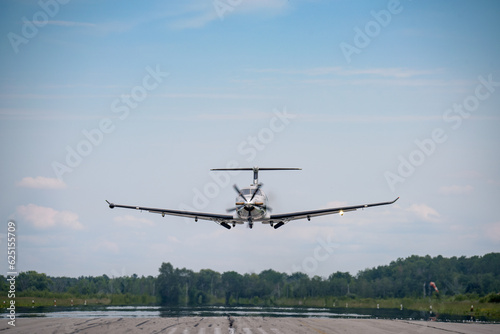 A small business turboprop Pilatus PC-12 on final with the landing gear starting to extend.  Blue sky with haze and clouds in the background.