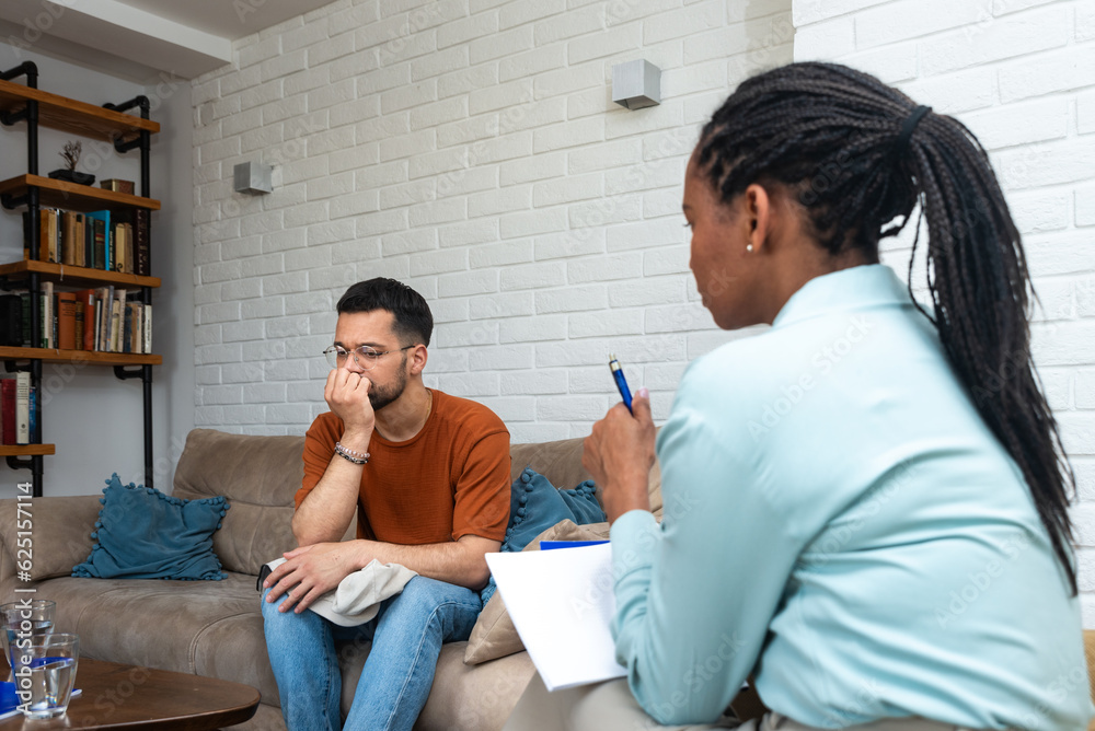 Young depressed man crying talking to mental health counselor during a session in the office. Stressed male at psychotherapy after divorce having emotional damage issue. Relationship difficulties.