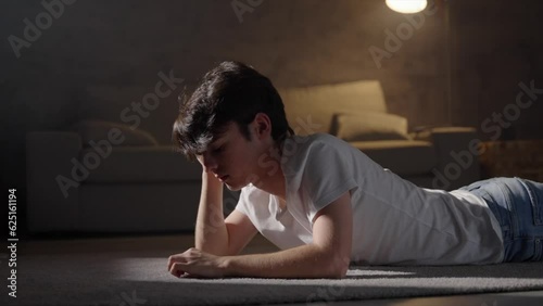 Sad teenager boy thinking lying on the floor at night. Worried person in the living room. Depression, loneliness and mental health concept.