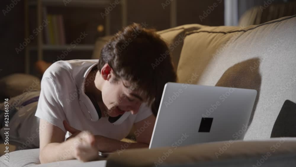 Teenager boy talking on laptop lying on sofa. Person having video call or live streaming at night.