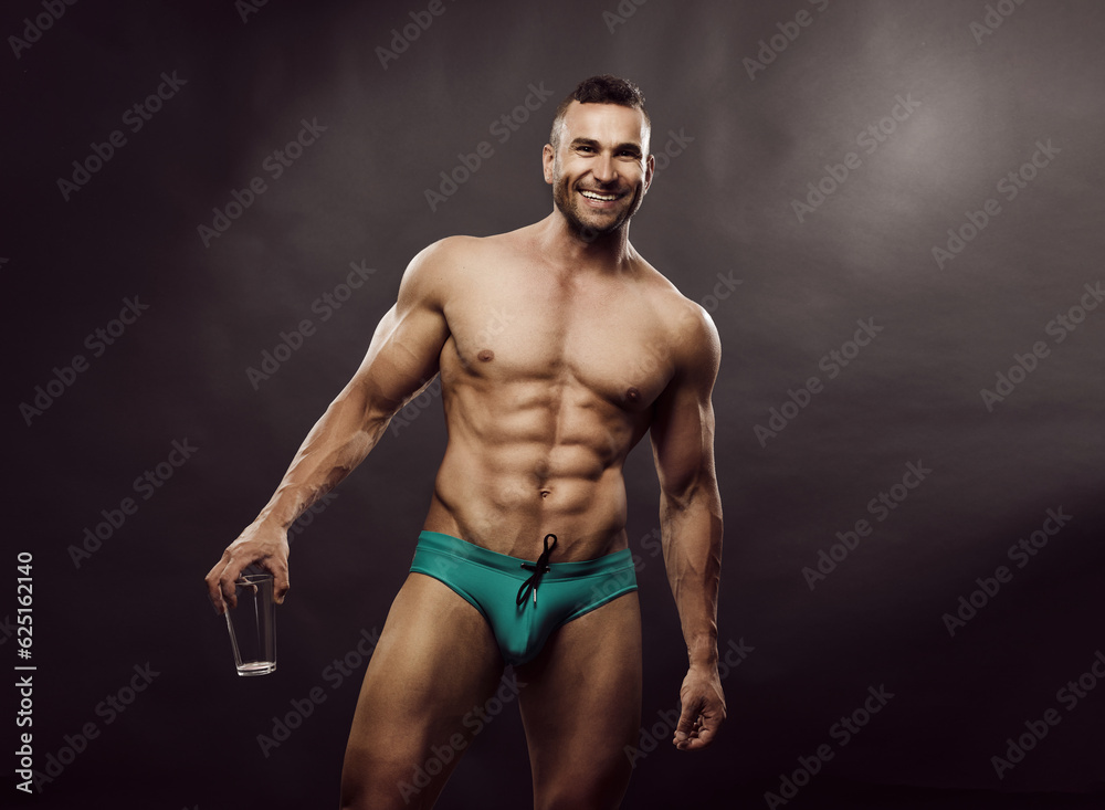 Tanned sports man in turquoise swimming trunks with glass in his hands. Smiling male model posing in the studio. Sexy bodybuilder with six pack abs in underwear in studio.