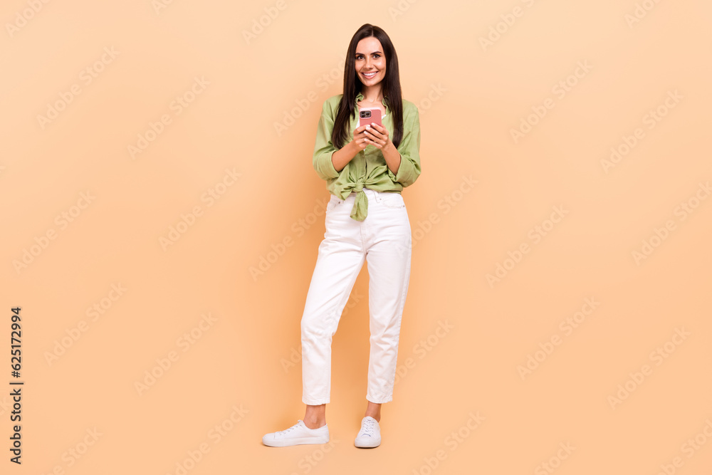 Full size photo of satisfied adorable nice girl dressed green blouse white trousers hold smartphone isolated on beige color background Full size photo of satisfied adorable nice girl dressed green blouse white trousers hold smartphone isolated on beige color background