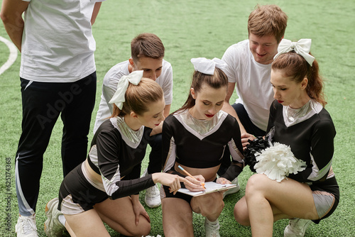Girl from cheerleader team making notes on paper and planning performance together with her team outdoors