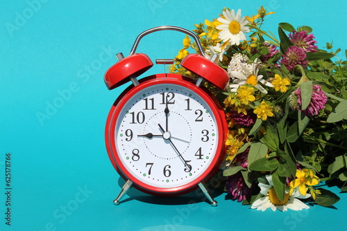 A red alarm clock stands on a turquoise background with a bouquet of wildflowers.