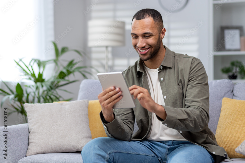 Hispanic smiling young man using tablet at home on sofa