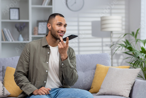 Fototapet African american young man sitting on sofa at home and talking on speaker phone,