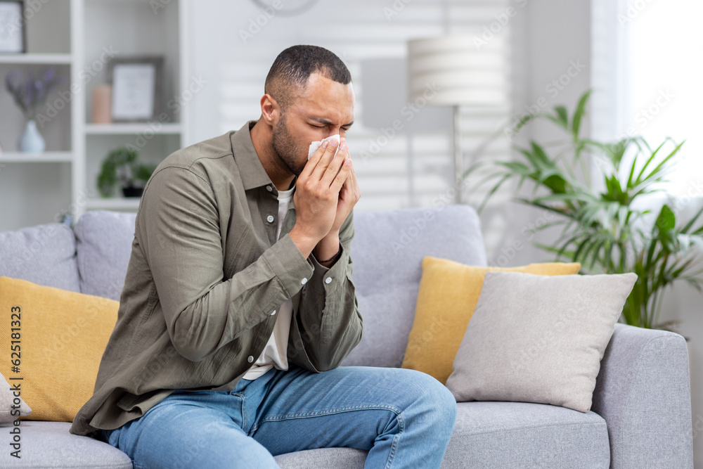 African American man sick at home. He sits on the couch and blows his nose into a tissue