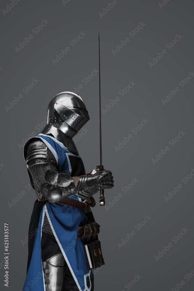 Naklejka premium A medieval soldier garbed in blue surcoat and armor holding a sword with a still, statue-like pose, against a gray background