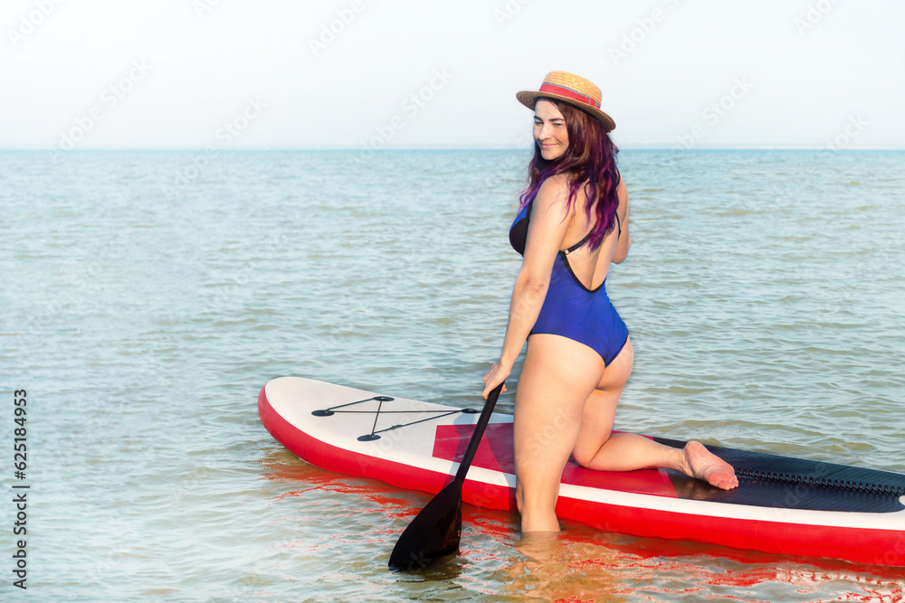 Sup boarding. A woman in a hat and blue swinsuit poses standing with a sup board. Back view. The concept of active lifestyle and surfing.