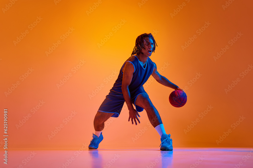 Young muscular man, athlete in motion, playing basketball, doing dribbling against orange background in neon lights. Concept of professional sport, competition, hobby, game, competition, ad