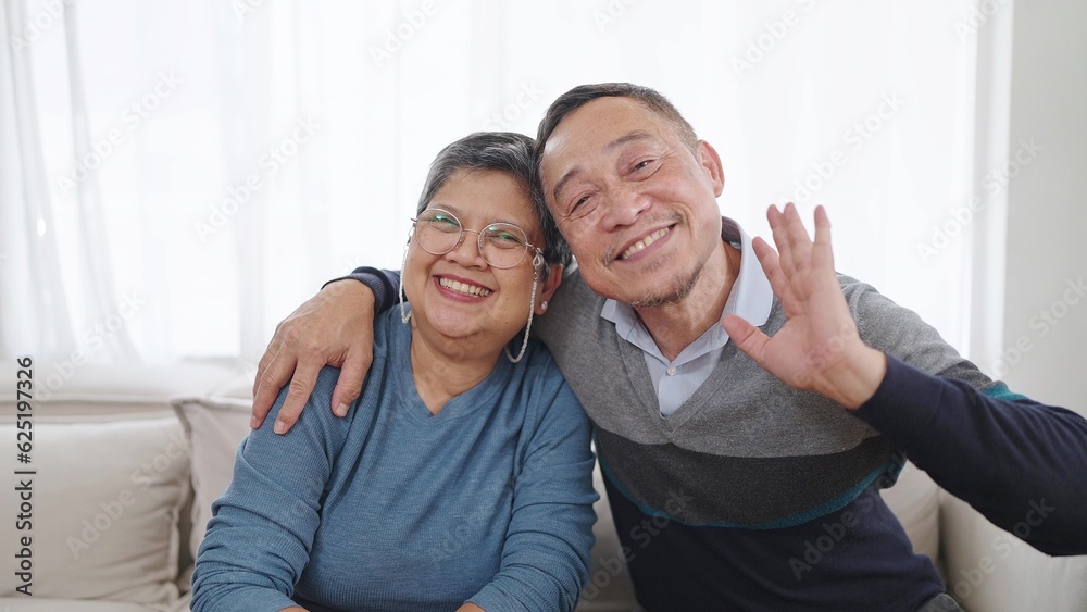 Happy elderly asian couple embracing bonding smiling and waving hands to camera while sitting on comfortable sofa in living room at home. Couple retirement lifestyle Happy elderly asian couple embracing bonding smiling and waving hands to camera while sitting on comfortable sofa in living room at home. Couple retirement lifestyle