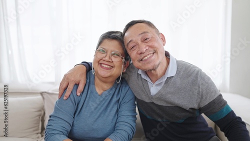 Happy elderly asian couple embracing bonding smiling and looking at camera while sitting on comfortable sofa in living room at home. Couple retirement lifestyle