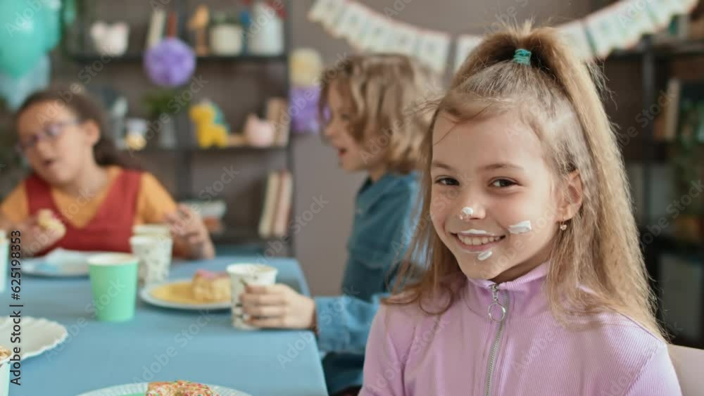 Medium close-up portrait shot of 9-year-old Caucasian girl sitting at table with friends at kids birthday party, enjoying sweets, looking at camera with cake cream on face and smiling Medium close-up portrait shot of 9-year-old Caucasian girl sitting at table with friends at kids birthday party, enjoying sweets, looking at camera with cake cream on face and smiling