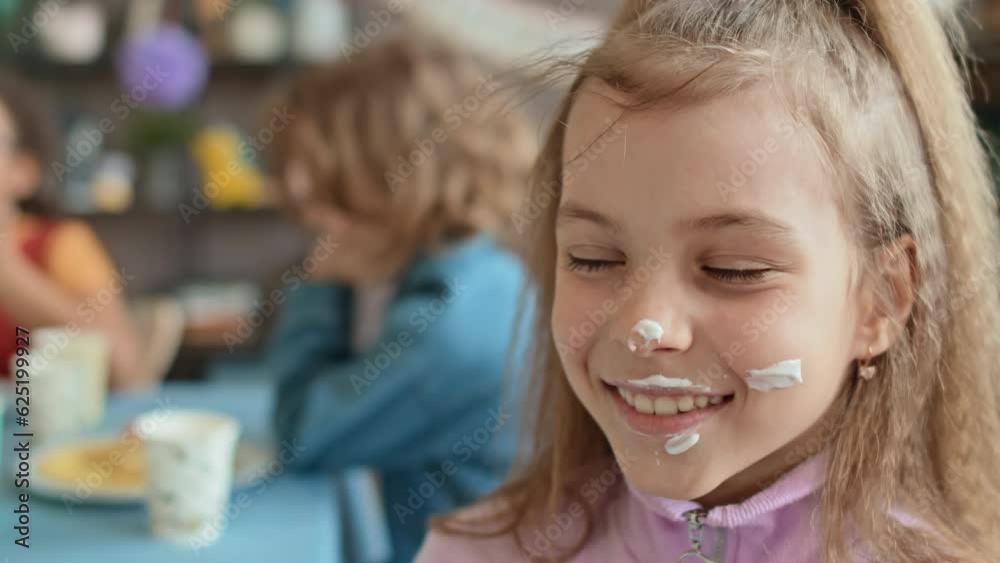 Close-up selective focus portrait shot of face of preteen Caucasian girl sitting at table with friends at kids birthday party, looking at camera with cake cream on face and smiling