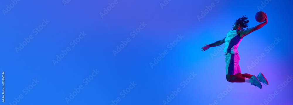 Professional basketball player in a jump with ball against gradient blue background in neon lights. Concept of professional sport, competition, hobby, game, competition. Banner. Copy space for ad