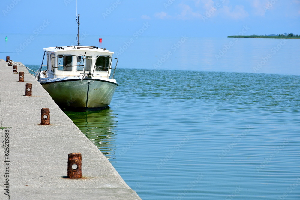 A close up on a small passenger or fishing boat parked next to a concrete part of a marina or jetty, next to some small channel covered with reeds, trees and other flors spotted in Poland in summer A close up on a small passenger or fishing boat parked next to a concrete part of a marina or jetty, next to some small channel covered with reeds, trees and other flors spotted in Poland in summer