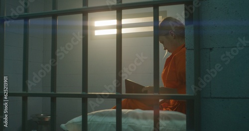 Photography Female prisoner in orange uniform sits on bed behind metal bars, reads Bible in prison cell, looks at barred window
