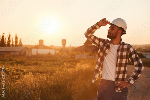 Touching the hard hat. Man is outdoors against sunset light. Rural scene