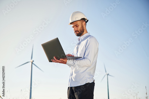 Standing and using laptop. Service engineer is on the field with windmills