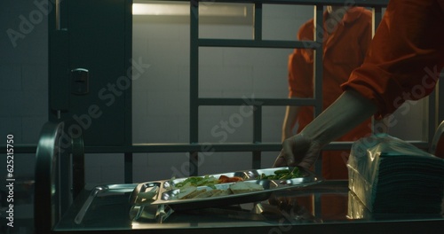 Prison worker gives food from serving trolley to female prisoner in orange uniform in jail cell. Woman criminal sits on bed, eats dinner. Imprisonment term in prison or detention center. Dolly shot.