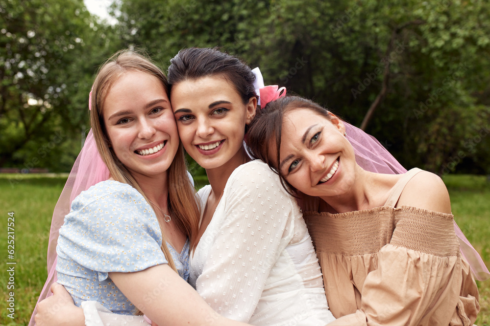 Happy adorable bride in a white dress with white veil and two bridesmaids in blue and beige dress with pink veils sitting on the grass in a summer park hugging each other. Cheerful bachelorette party. Happy adorable bride in a white dress with white veil and two bridesmaids in blue and beige dress with pink veils sitting on the grass in a summer park hugging each other. Cheerful bachelorette party.