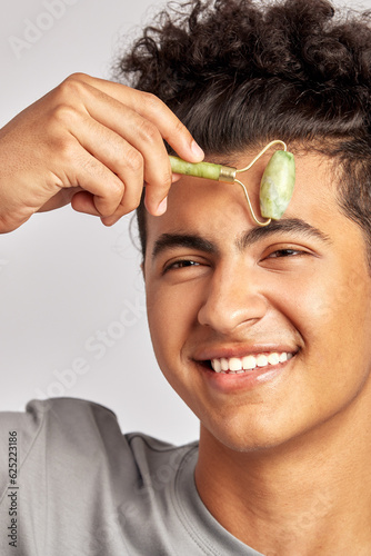 Young handsome smiling guy using jade de-puffing face roller to keep his skin soft and smooth. Swarthy man with black curly hair maintains his natural beauty with a skincare routine. Men's self-care.