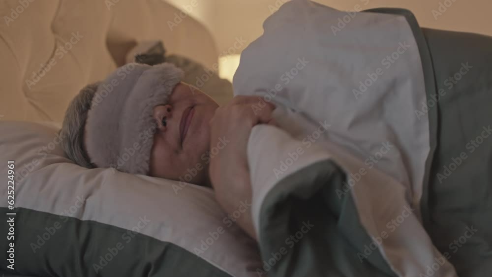 Peaceful senior Asian woman lying on pillow under soft blanket, putting on silk sleeping mask and falling asleep in cozy bed at night Peaceful senior Asian woman lying on pillow under soft blanket, putting on silk sleeping mask and falling asleep in cozy bed at night