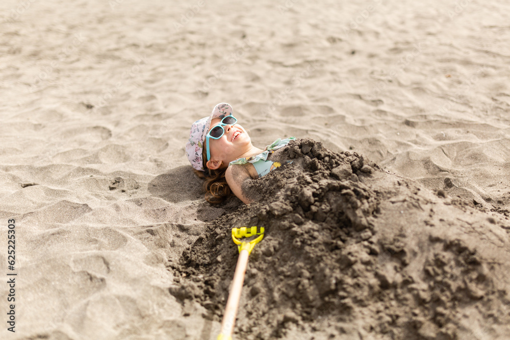 Happy kid in cap and sunglasses lying on ground with wet sand on body Happy kid in cap and sunglasses lying on ground with wet sand on body