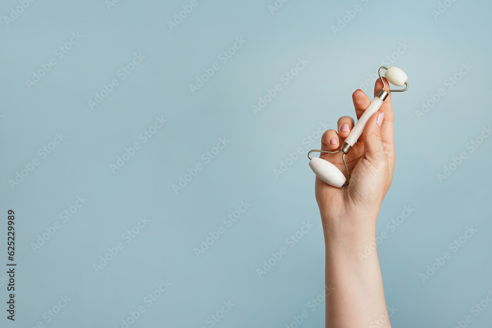 Hand holding white facial jade massage roller, gua sha on blue background. Skin care, copy space Hand holding white facial jade massage roller, gua sha on blue background. Skin care, copy space