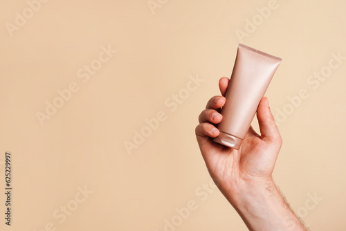Male hand holding unbranded beige tube with lotion or cosmetics cream on yellow background. Shaving foam or male skin care concept. Minimalism, copy space