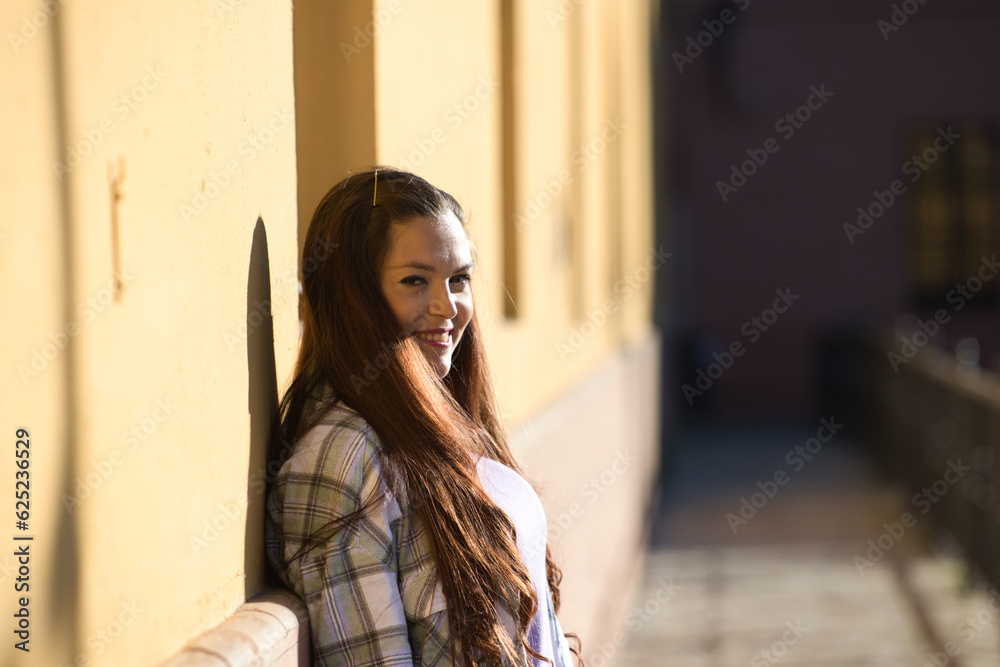Beautiful young Spanish woman leaning against yellow wall. The woman is happy and enjoying the sunny day.