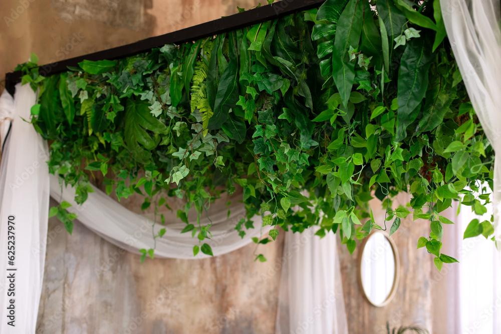 Composition from garland flowers and plants on wall. Ceiling decoration. Wall with curly green plants garlands hanging from ceiling. Herb and plant wall, nature wall. Interior wedding party decor. - obrazy, fototapety, plakaty 