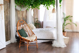 Interior boho design bedroom with garland flowers and plants. Cozy light bedroom in Scandinavian style bedroom, canopy bed and  wicker rattan armchair. Bed decorated flowing white curtains canopies. 