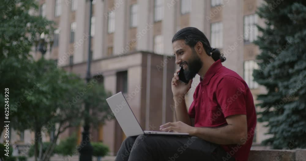 Portrait of Middle Eastern person working with laptop and discussing idea on mobile phone sitting outdoors in city. Communication and portable devices concept. Portrait of Middle Eastern person working with laptop and discussing idea on mobile phone sitting outdoors in city. Communication and portable devices concept.