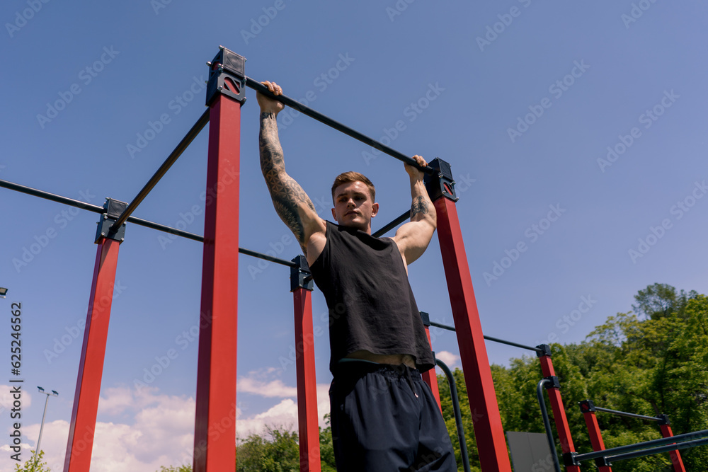 Fototapeta premium portrait of a young sweaty sportsman doing horizontal bar swings arms and shoulders push-ups on the sports ground during street training