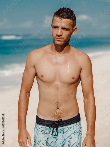 Handsome young muscular man posing shirtless on ocean beach, summer time. Ideal slim fit body.