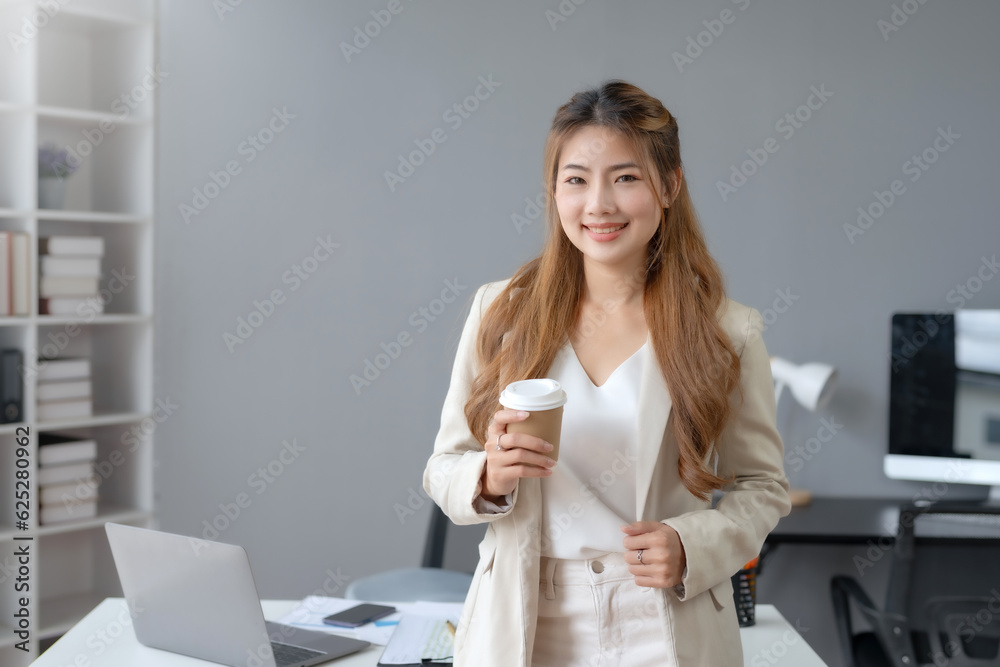 Successful Asian businesswoman smiling using laptop computer and holding coffee cup at office. Confident Asia businesswoman sitting happily in the office. Successful Asian businesswoman smiling using laptop computer and holding coffee cup at office. Confident Asia businesswoman sitting happily in the office.