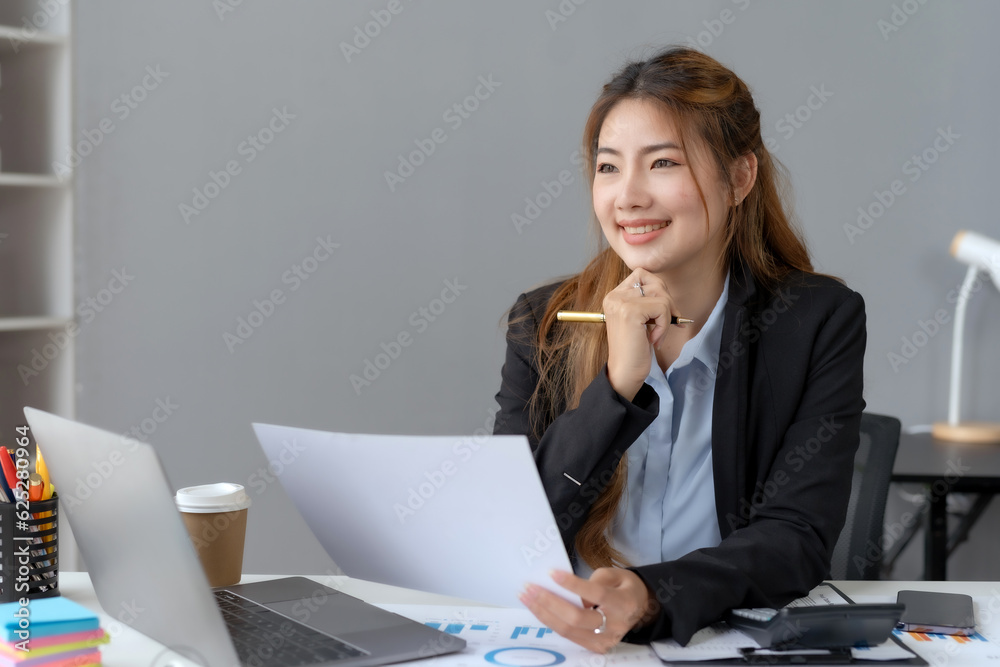 Happy Asian woman accountant at work sitting and taking note of the office report. Asian businesswoman writing a note making a report on investment profits earnings of a company. Happy Asian woman accountant at work sitting and taking note of the office report. Asian businesswoman writing a note making a report on investment profits earnings of a company.